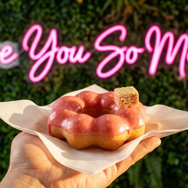 Hand holding a pink glazed mochi donut with a wafer, in front of a neon "Love You So Mochi" sign.